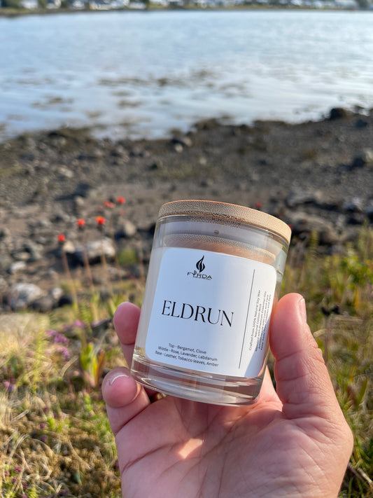 Hand holding Eldrun soy candle by Fyrda near rocky coastal shoreline with wildflowers in sunlight