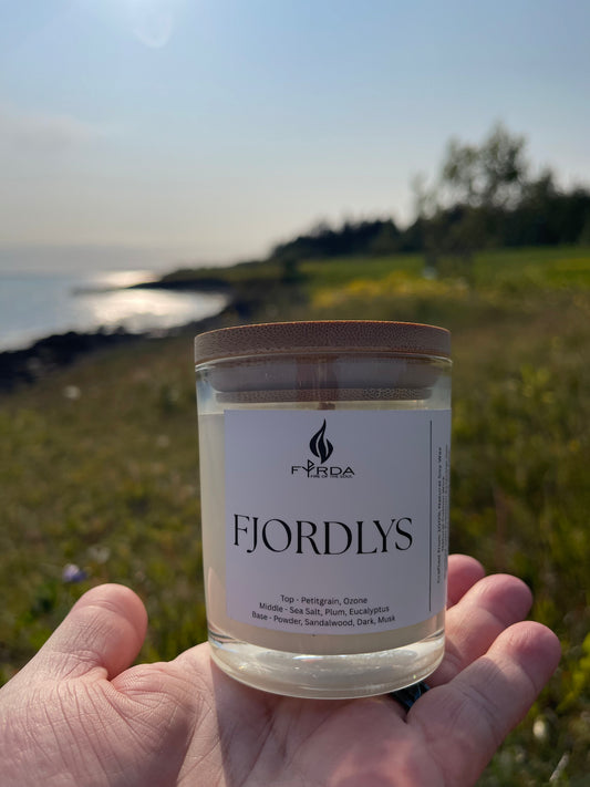Hand holding FJORDLYS candle outdoors near the coast during golden hour, glowing meadow and sunset sky in background