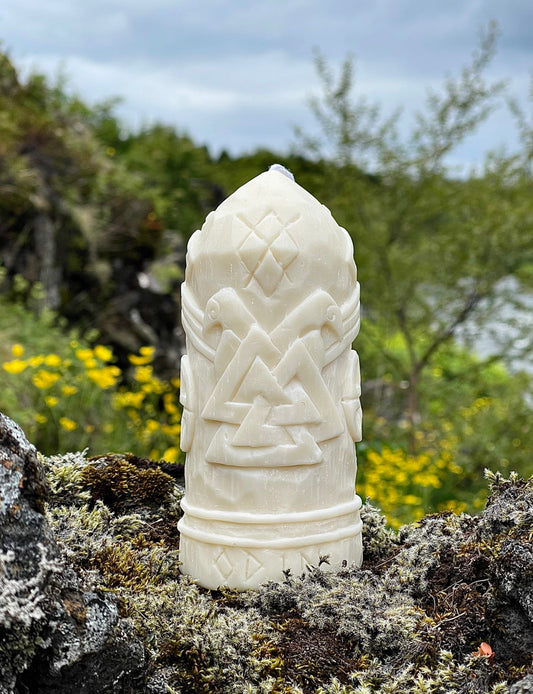 Odin candle with Valknut symbol and two ravens carved in soy wax, placed on mossy rock with wildflowers in the background