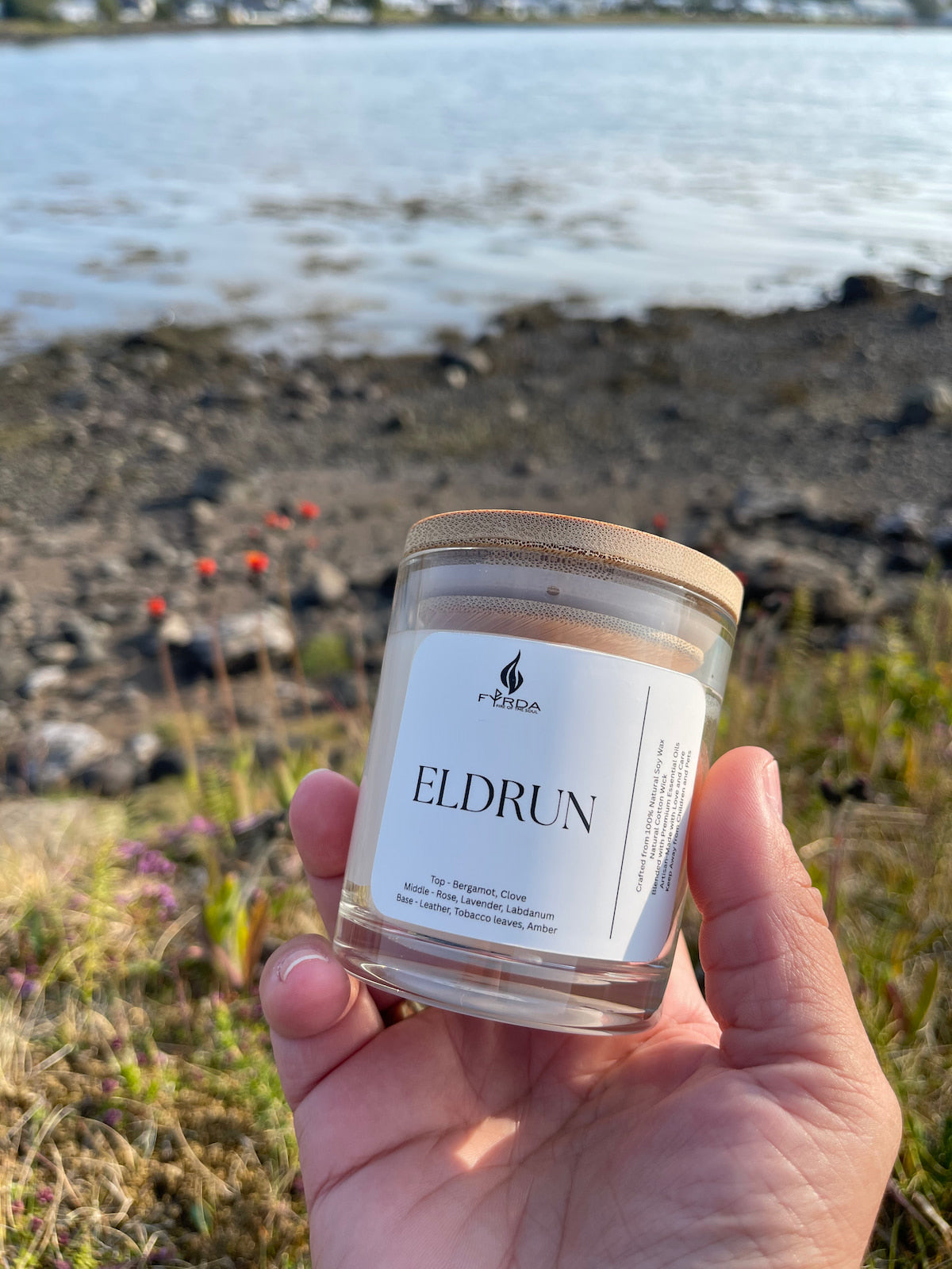 Hand holding Eldrun soy candle by Fyrda near rocky coastal shoreline with wildflowers in sunlight