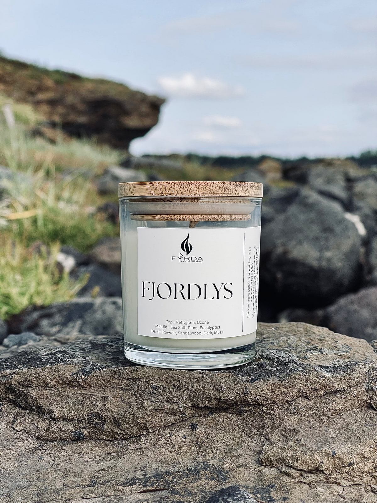FJORDLYS soy candle with wooden lid standing on coastal rock with cliff and blue sky in the background