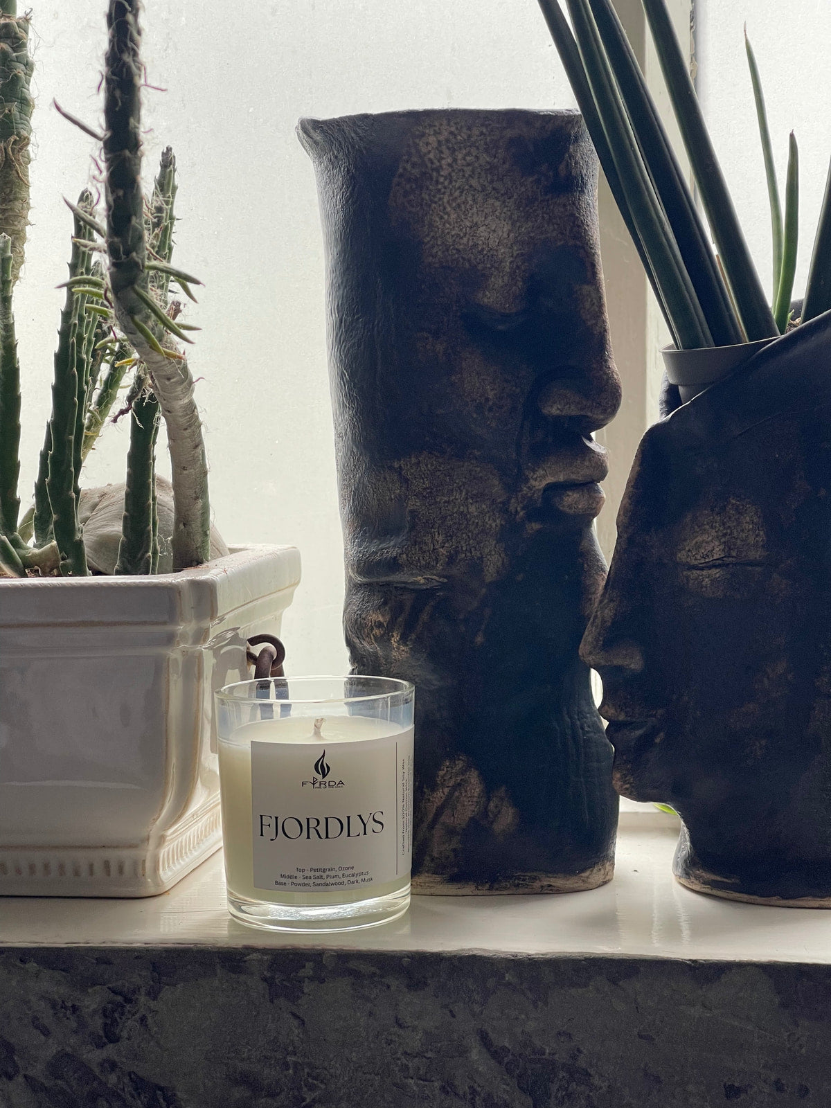 FJORDLYS soy candle on windowsill beside indoor plants and decorative ceramic face vases in soft natural light