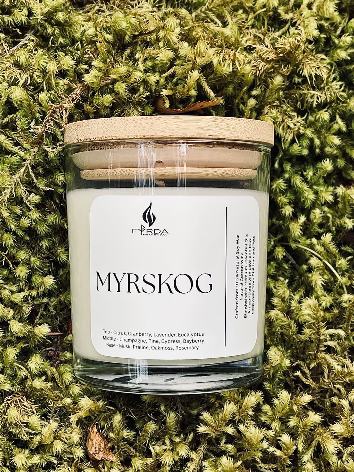 Hand holding MYRSKOG soy candle with wooden lid in front of rocky coastal shore and wildflowers, bright summer light