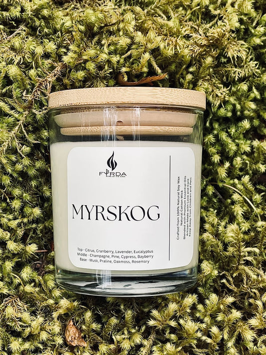 Hand holding MYRSKOG soy candle with wooden lid in front of rocky coastal shore and wildflowers, bright summer light
