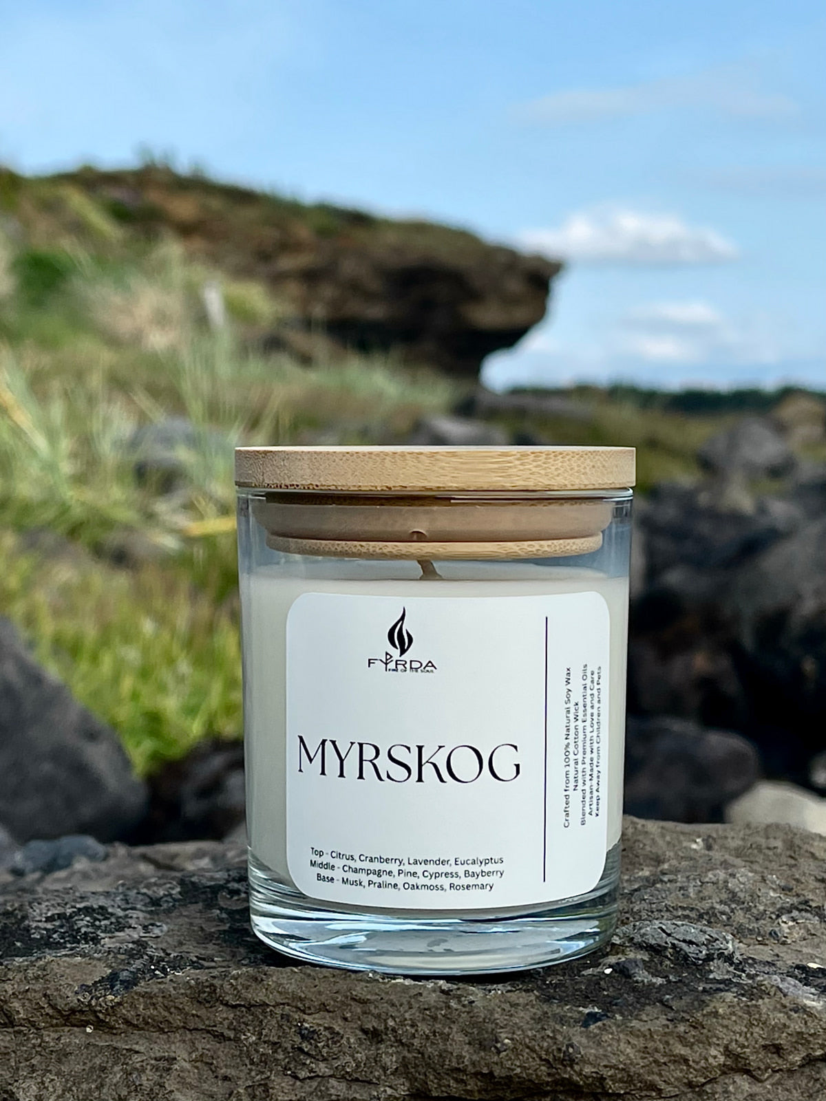 MYRSKOG soy candle with wooden lid placed on rugged rock, wild cliff and grassy background under blue sky
