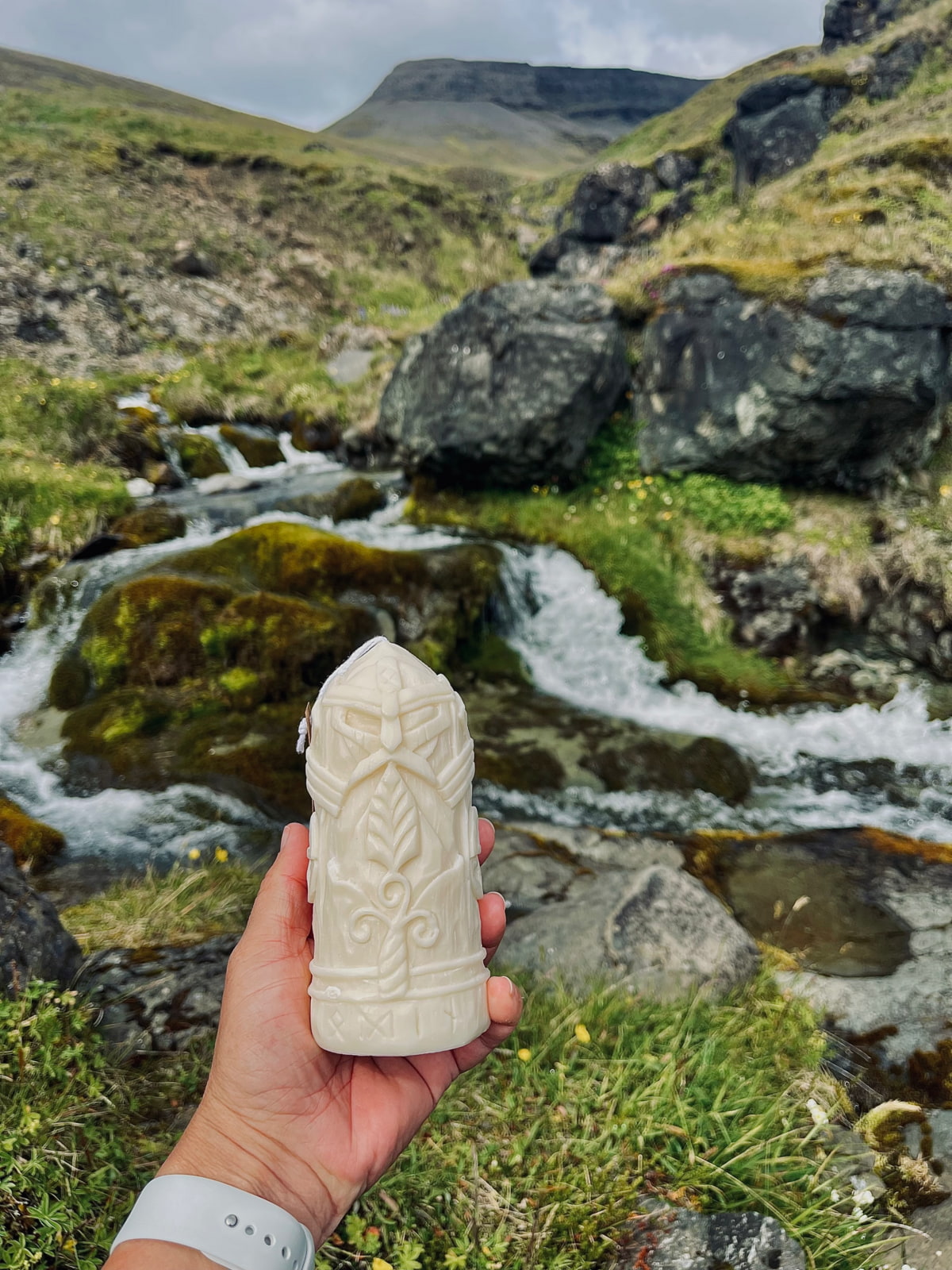 Hand holding Odin candle carved from soy wax, with Viking mask design, in front of Icelandic river and mossy mountain landscape