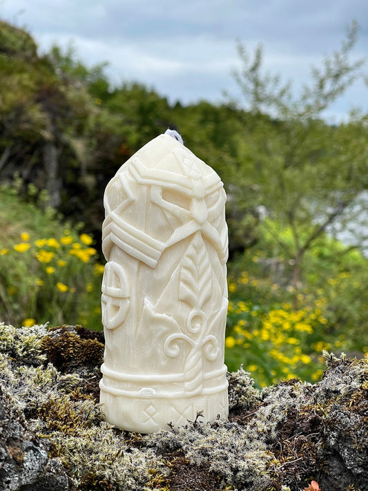 Hand-carved soy wax candle of Odin wearing a Viking mask, placed on mossy stone with wildflowers in the background